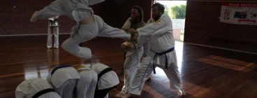 Young lady doing a flying kick over students and through a wooden board.